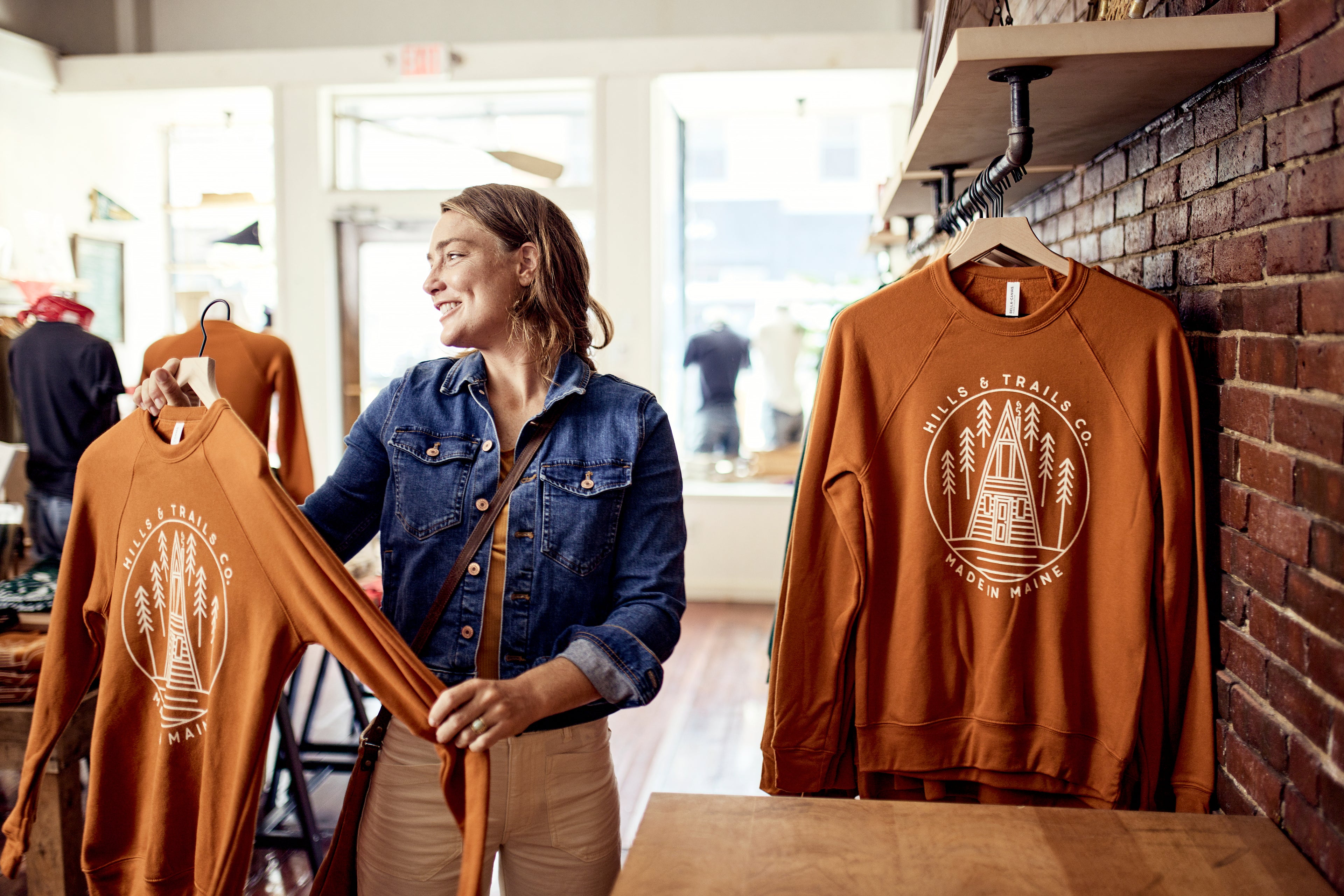 A smiling woman browses a shop, holding up a rust-colored “Hills & Trails Co. – Made in Maine” sweatshirt, with more apparel displayed around her as natural light filters through large windows.